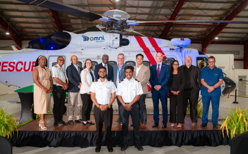 Group photo at the Kesi Adele Adams Memorial Heliport in Ogle, featuring the first two Guyanese ab-initio pilots, Somant Heeralall and Yadram Ramcharran, standing in front of an Omni Taxi Aéreo helicopter, alongside Omni Helicopters International executives, government officials, and guests celebrating the milestone training completion.